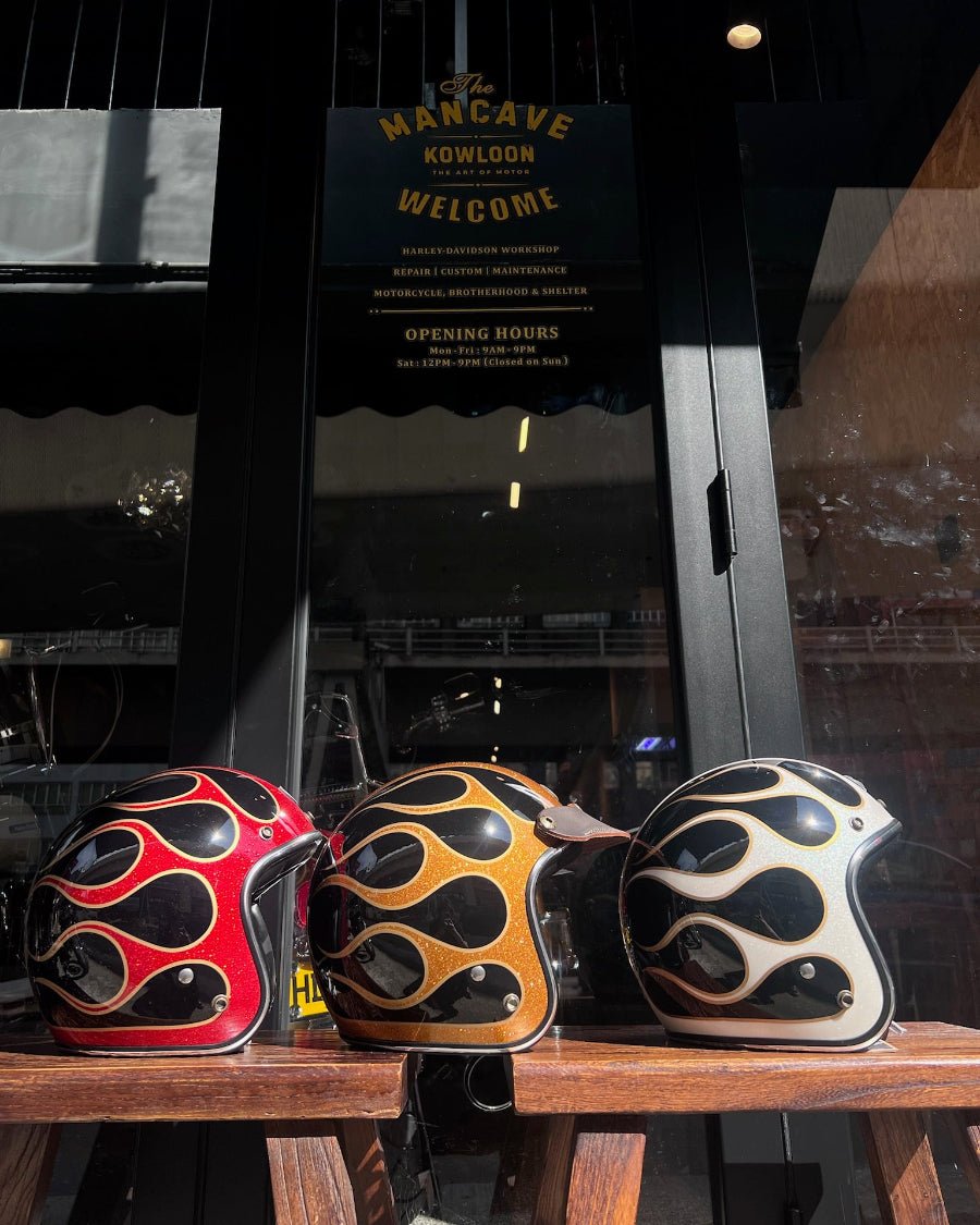 Three motorcycle helmets with flame design on a wooden shelf in front of a glass door with 'The Man Cave Kowloon' sign.