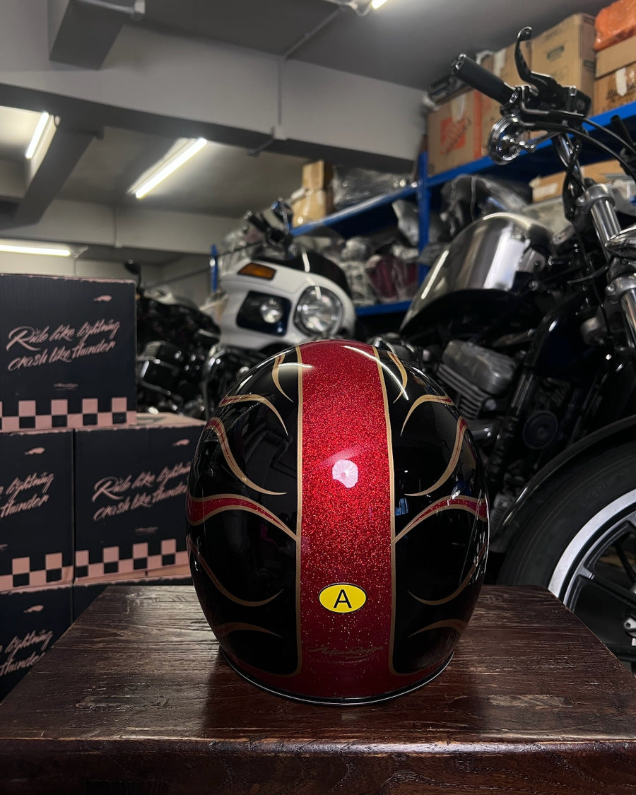 Red and black helmet with a yellow label on a wooden surface, with motorcycles and boxes in the background.