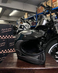 Black motorcycle helmet on a wooden surface with a motorcycle in the background.