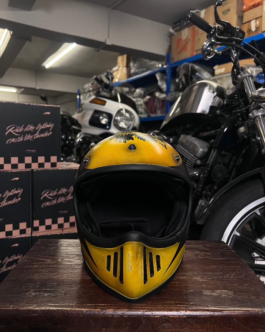Yellow motorcycle helmet on a wooden surface with a motorcycle and boxes in the background.