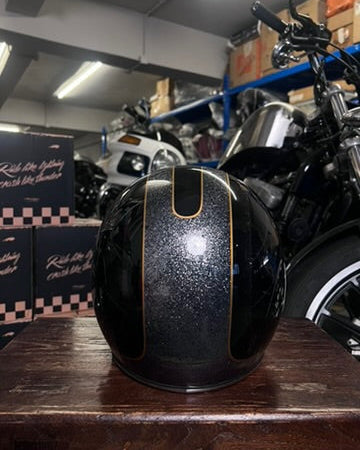 Black motorcycle helmet with gold accents on a wooden surface, with a motorcycle and boxes in the background.