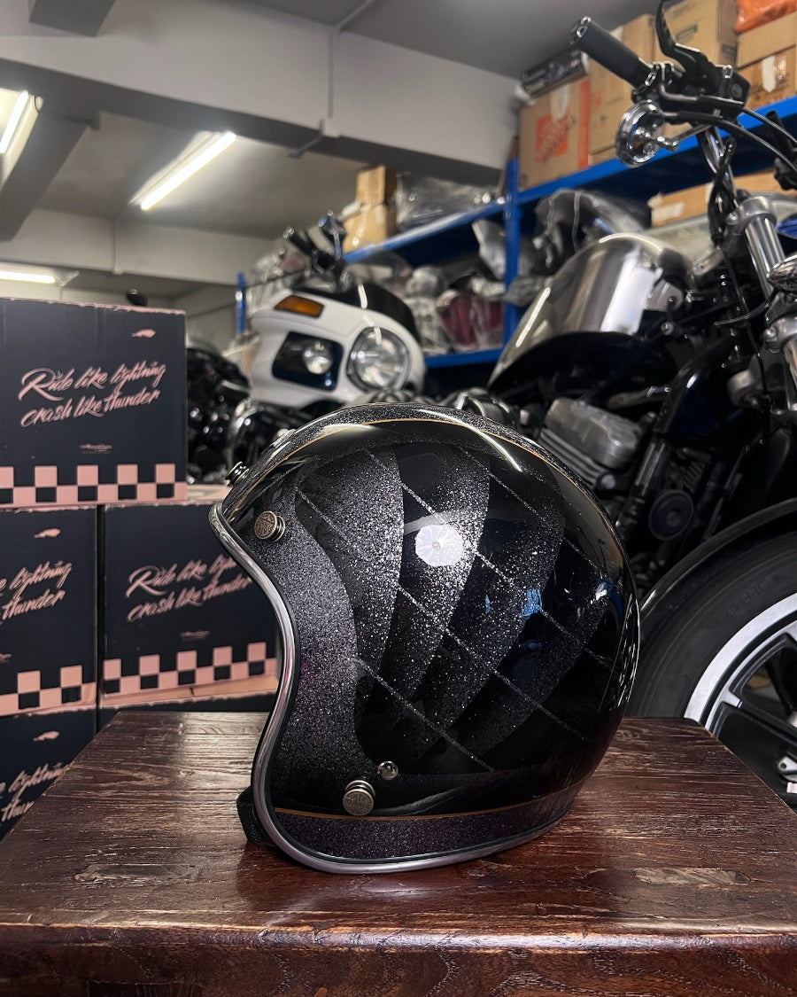 Black helmet with quilted design on a wooden surface in a garage setting with motorcycles and boxes.