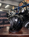 Black helmet with quilted design on a wooden surface in a garage setting with motorcycles and boxes.