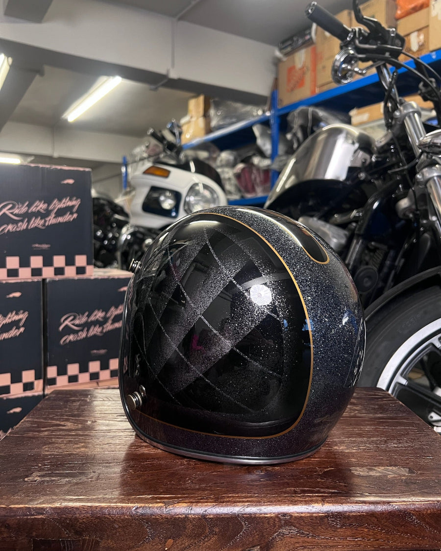 Black quilted motorcycle helmet on a wooden surface with a motorcycle and boxes in the background.