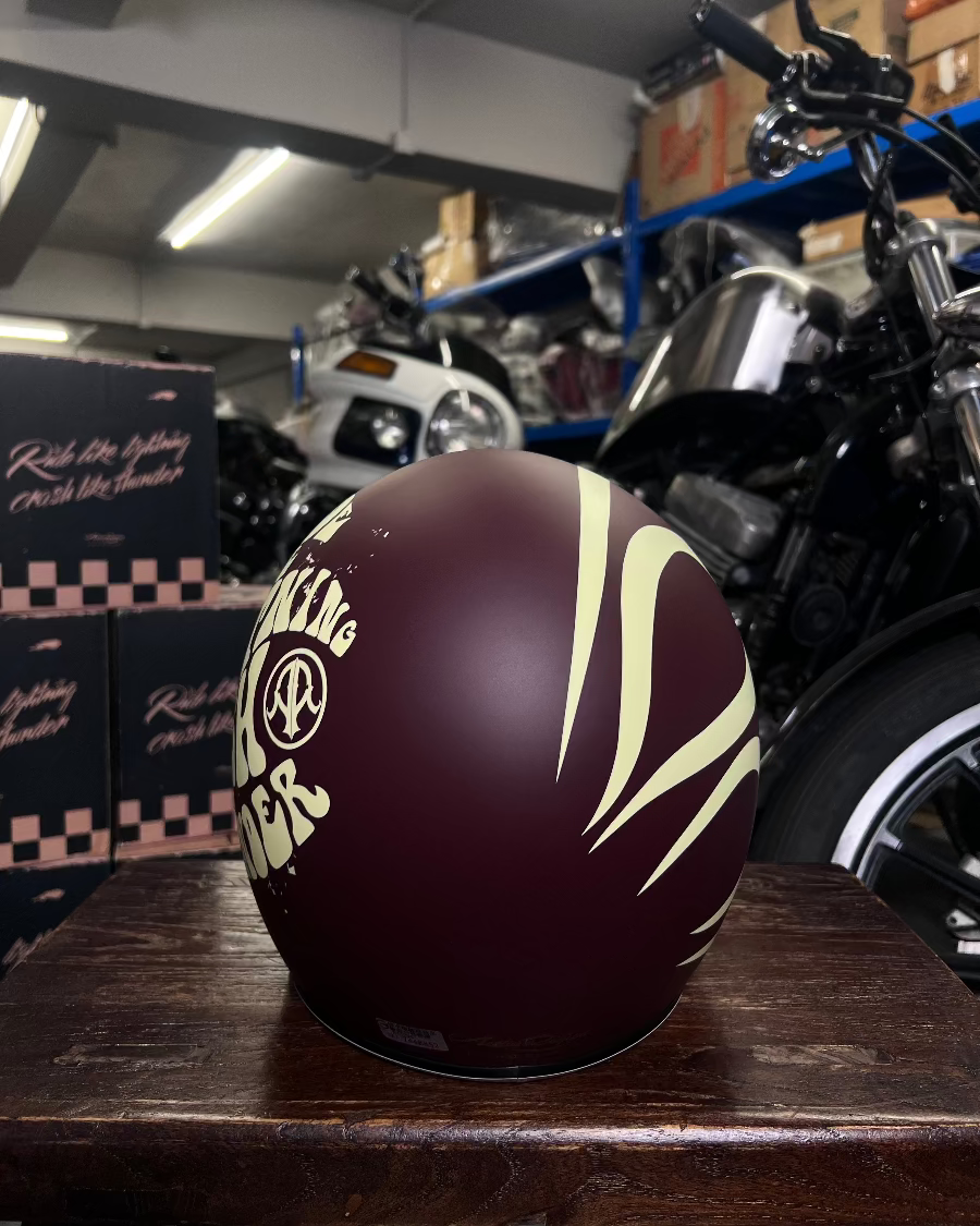 Maroon motorcycle helmet with white designs on a wooden surface, with a motorcycle and boxes in the background.