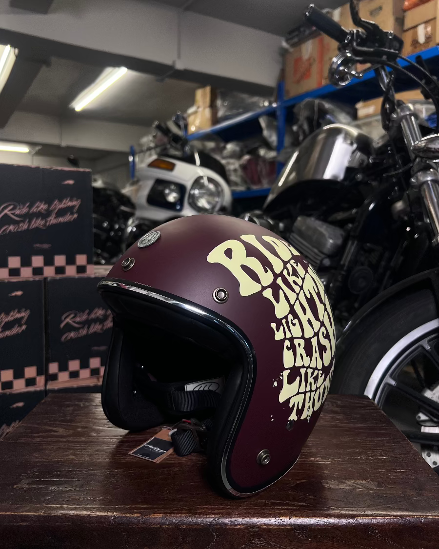 Maroon motorcycle helmet with yellow text on a wooden surface, with a motorcycle and boxes in the background.