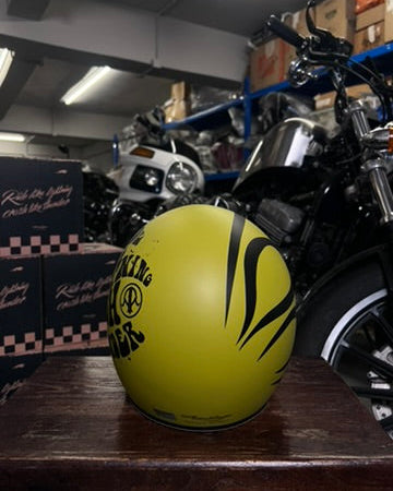 Yellow motorcycle helmet with black designs on a wooden surface, with a motorcycle and boxes in the background.