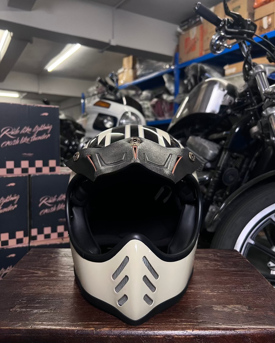 Black and beige motorcycle helmet on a wooden surface with a motorcycle and boxes in the background.