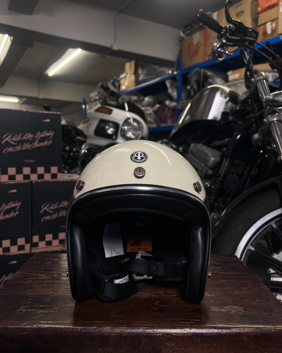 Beige motorcycle helmet with a logo on a wooden surface, with motorcycles and boxes in the background.
