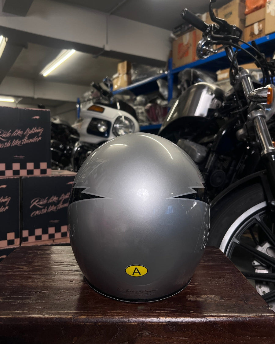 Gray motorcycle helmet with a yellow label on a wooden surface, with motorcycles and boxes in the background.