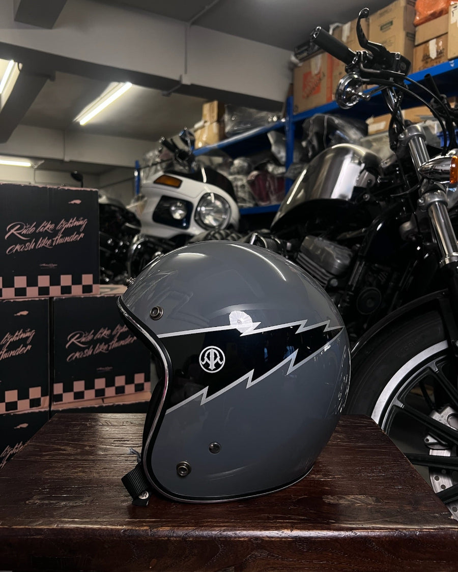 Motorcycle helmet with a black and gray design on a wooden surface, with a motorcycle and boxes in the background.